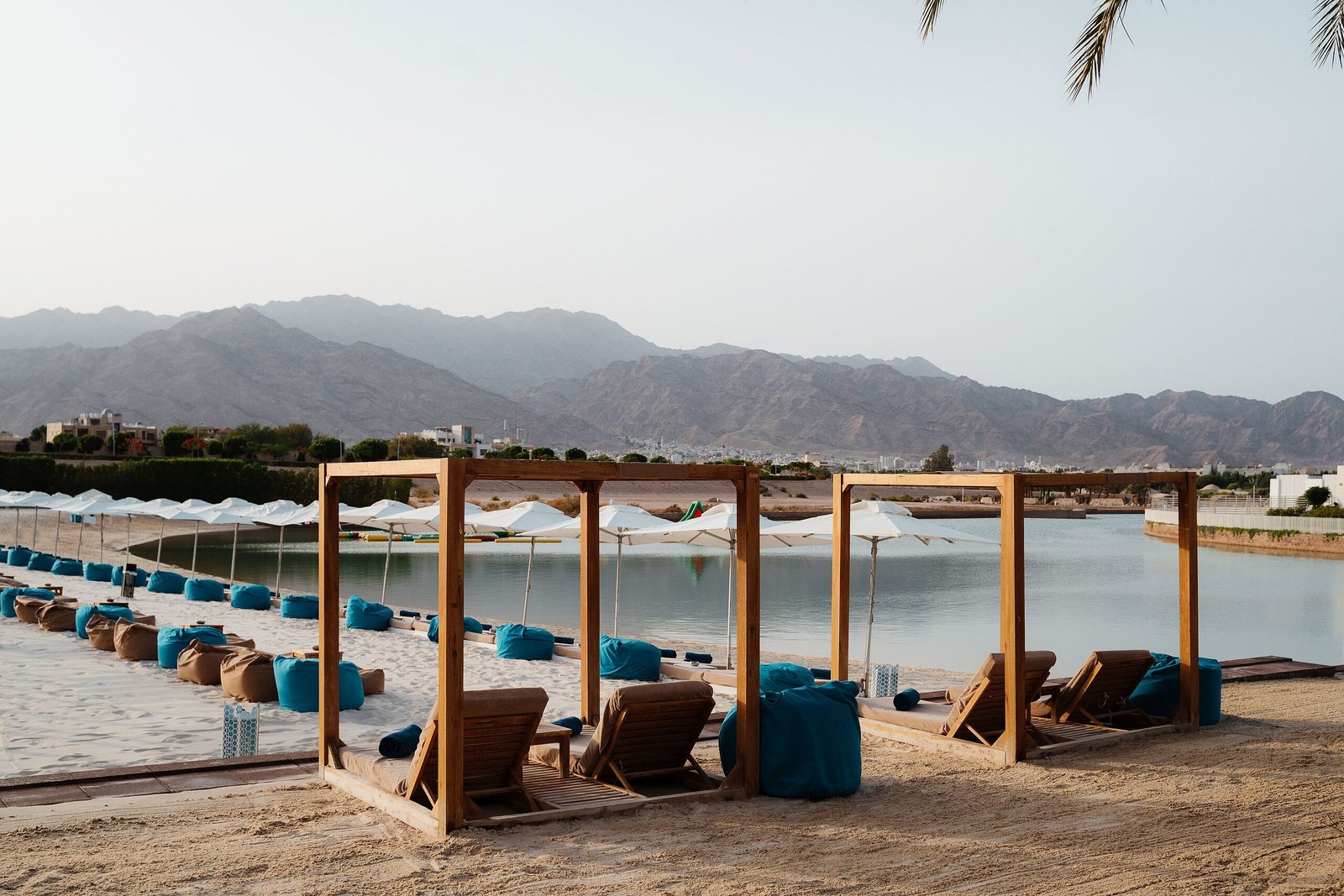 Row of wooden beach cabanas and loungers on a sand beach beside calm water, with mountains beyond, Aqaba.
