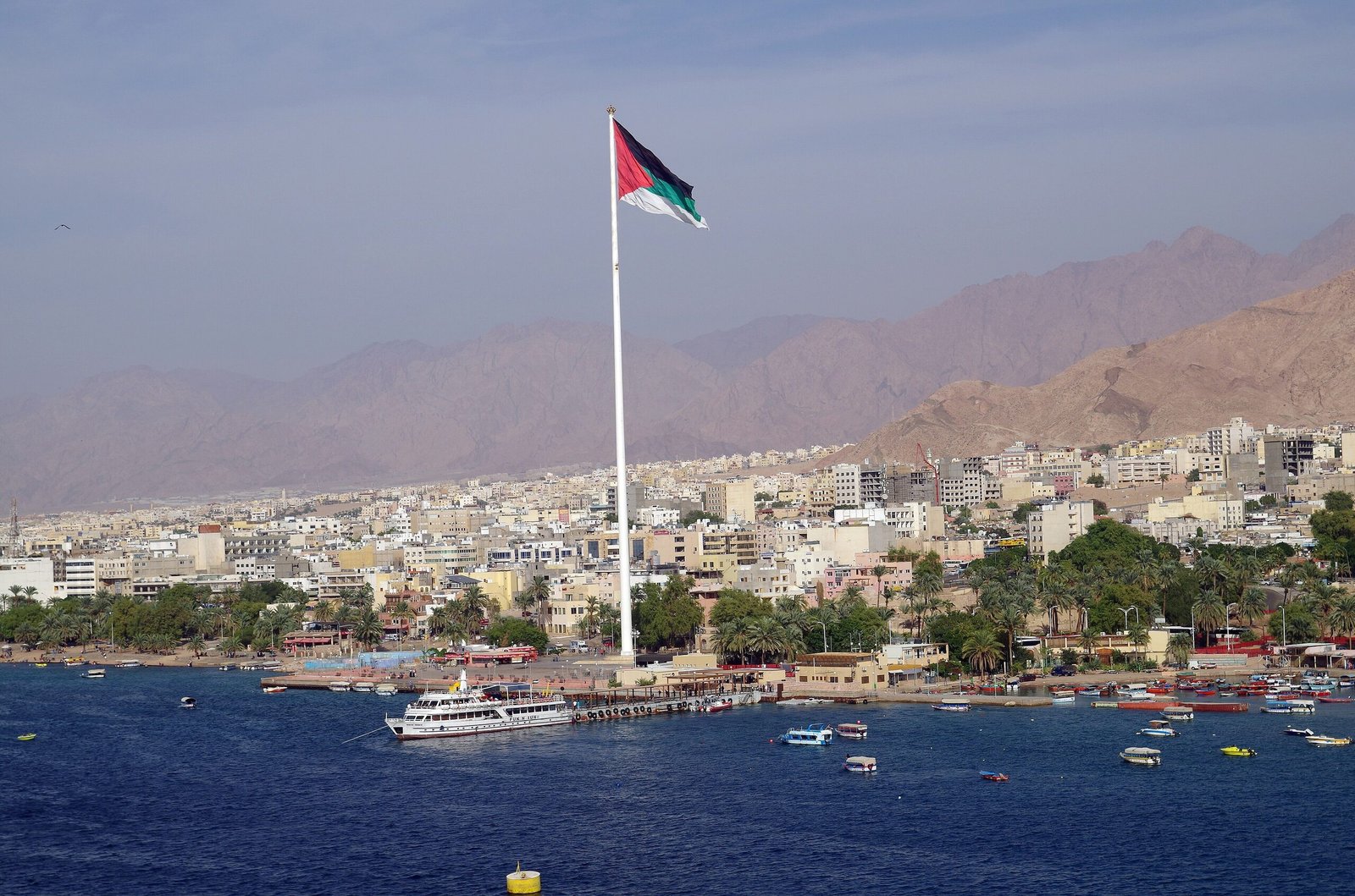 The Great Arab Revolt flag flying from its mast above the Aqaba waterfront, with the city and mountains behind.