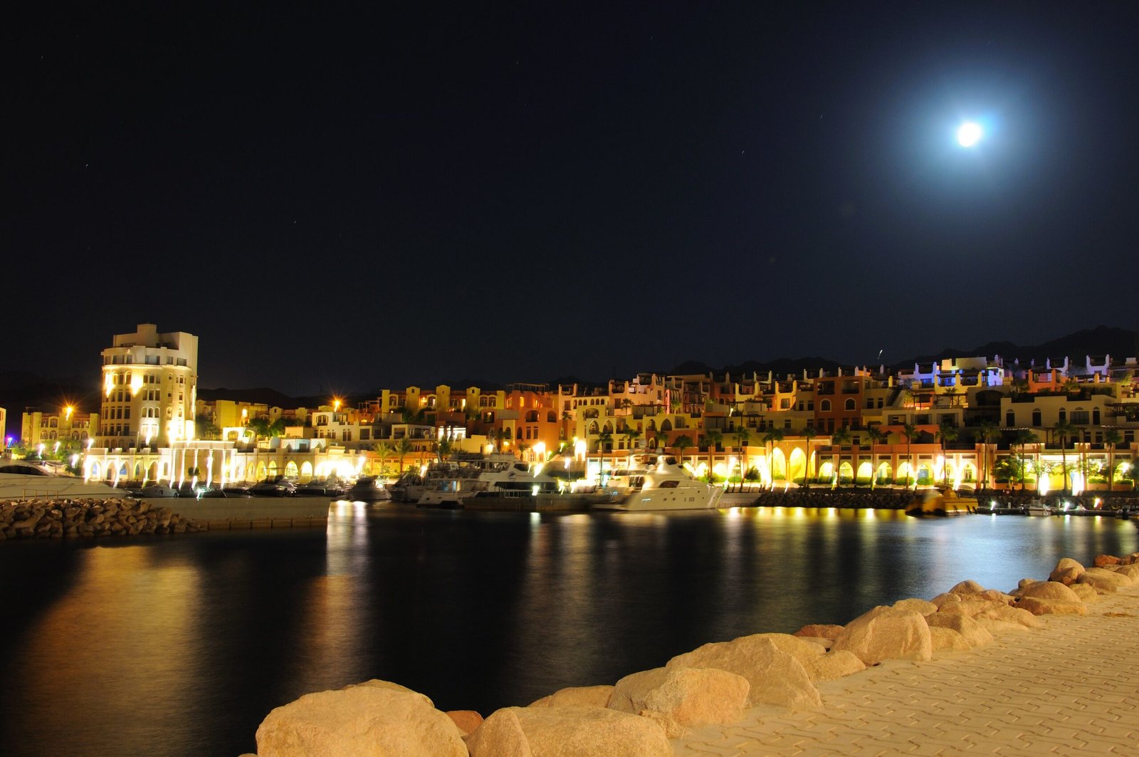Illuminated waterfront buildings of Tala Bay marina in Aqaba reflected in dark water at night.