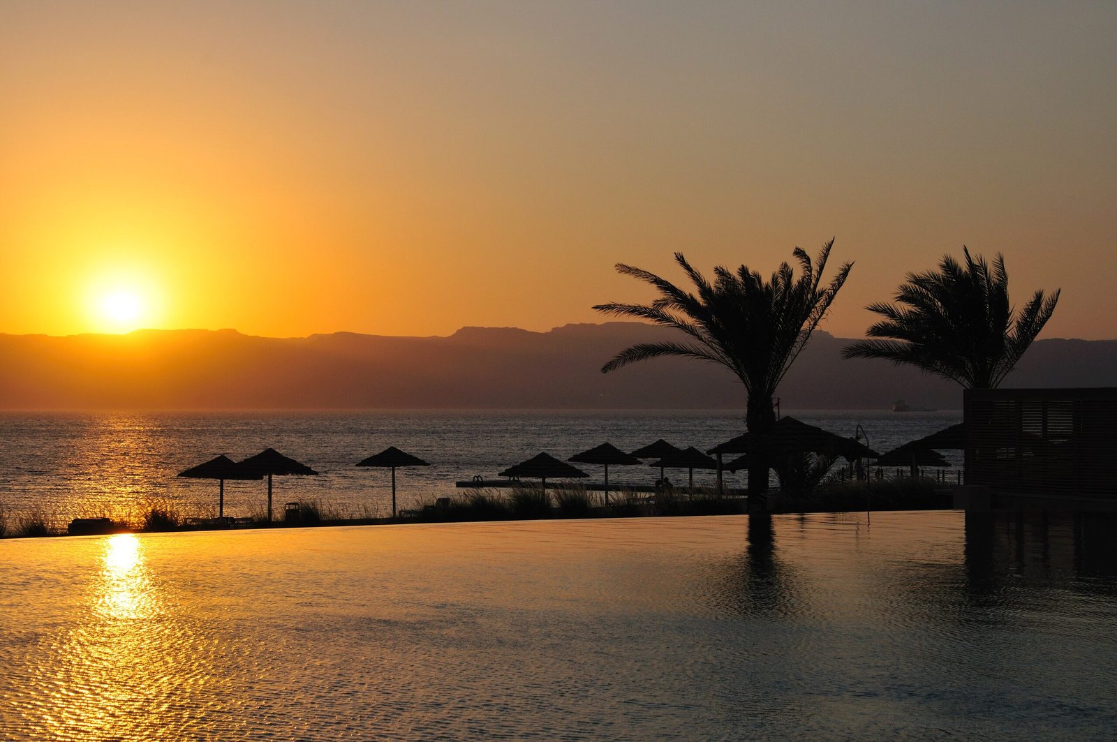 Sun setting over the Gulf of Aqaba behind mountains, seen past thatched parasols and palms at Tala Bay.