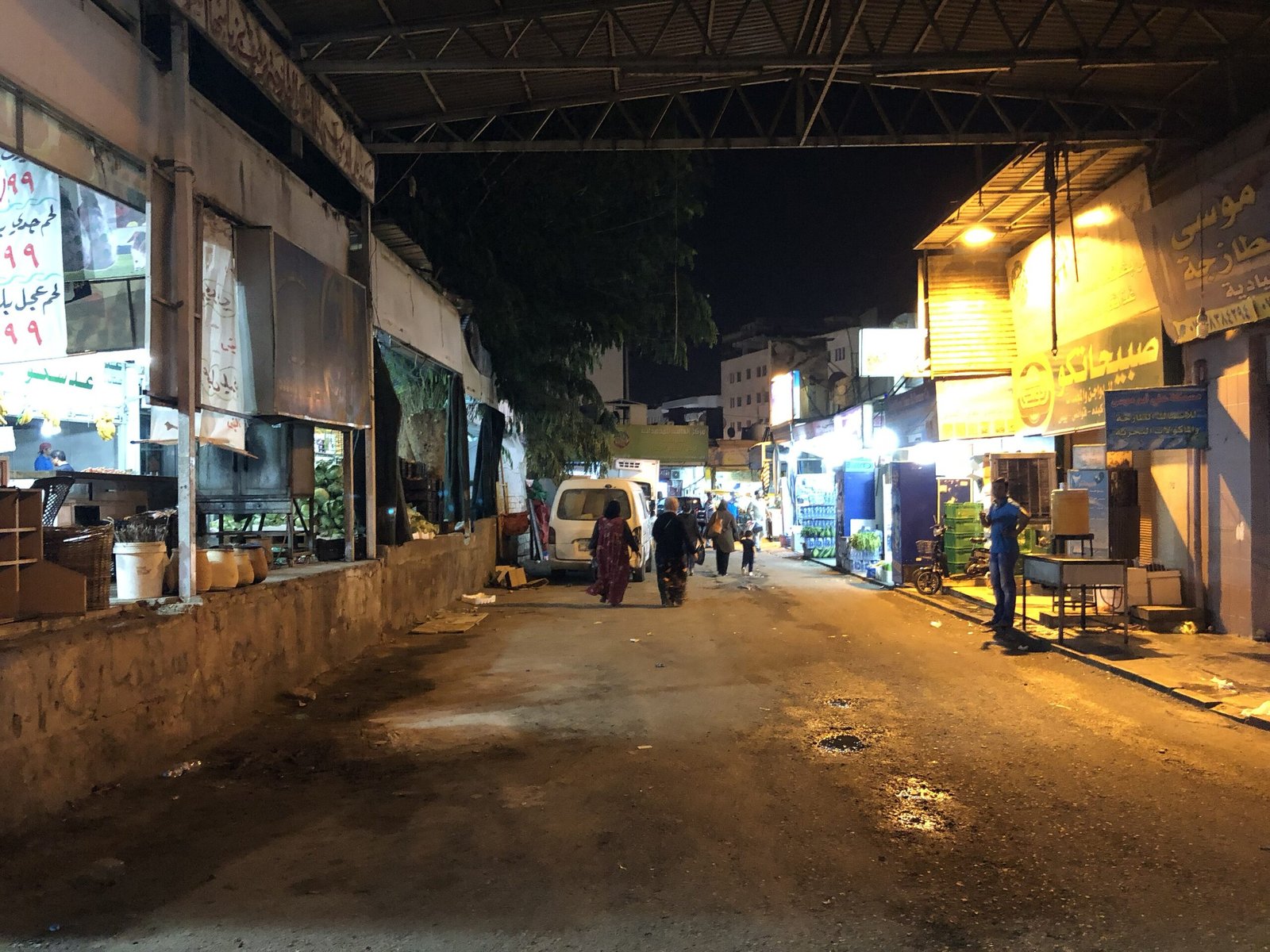 A lane in the Aqaba market district after dark, shopfronts lit on both sides and people walking between them.