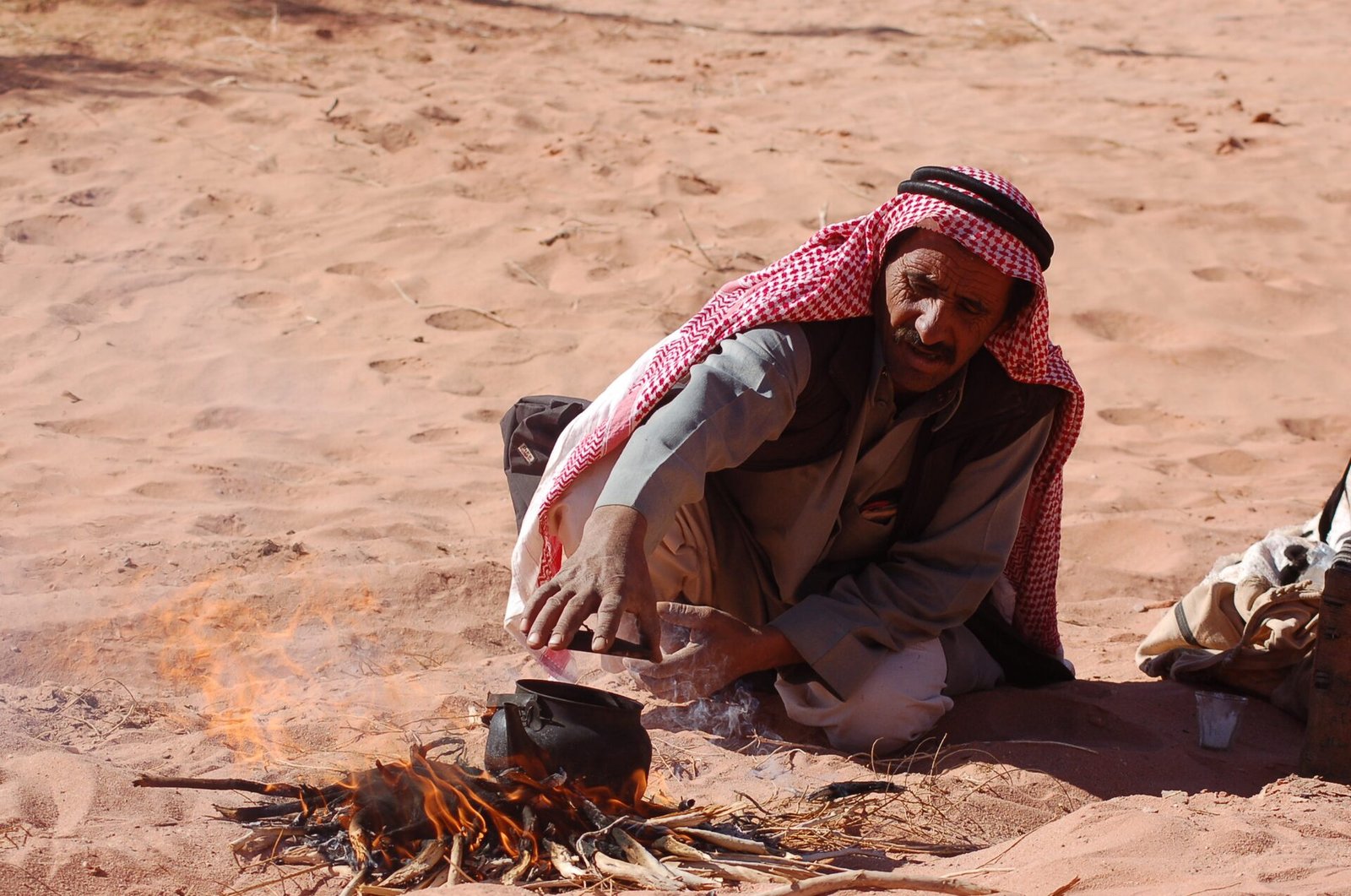 Bedouin man in a red keffiyeh crouching to make tea over an open fire on desert sand in Wadi Rum.