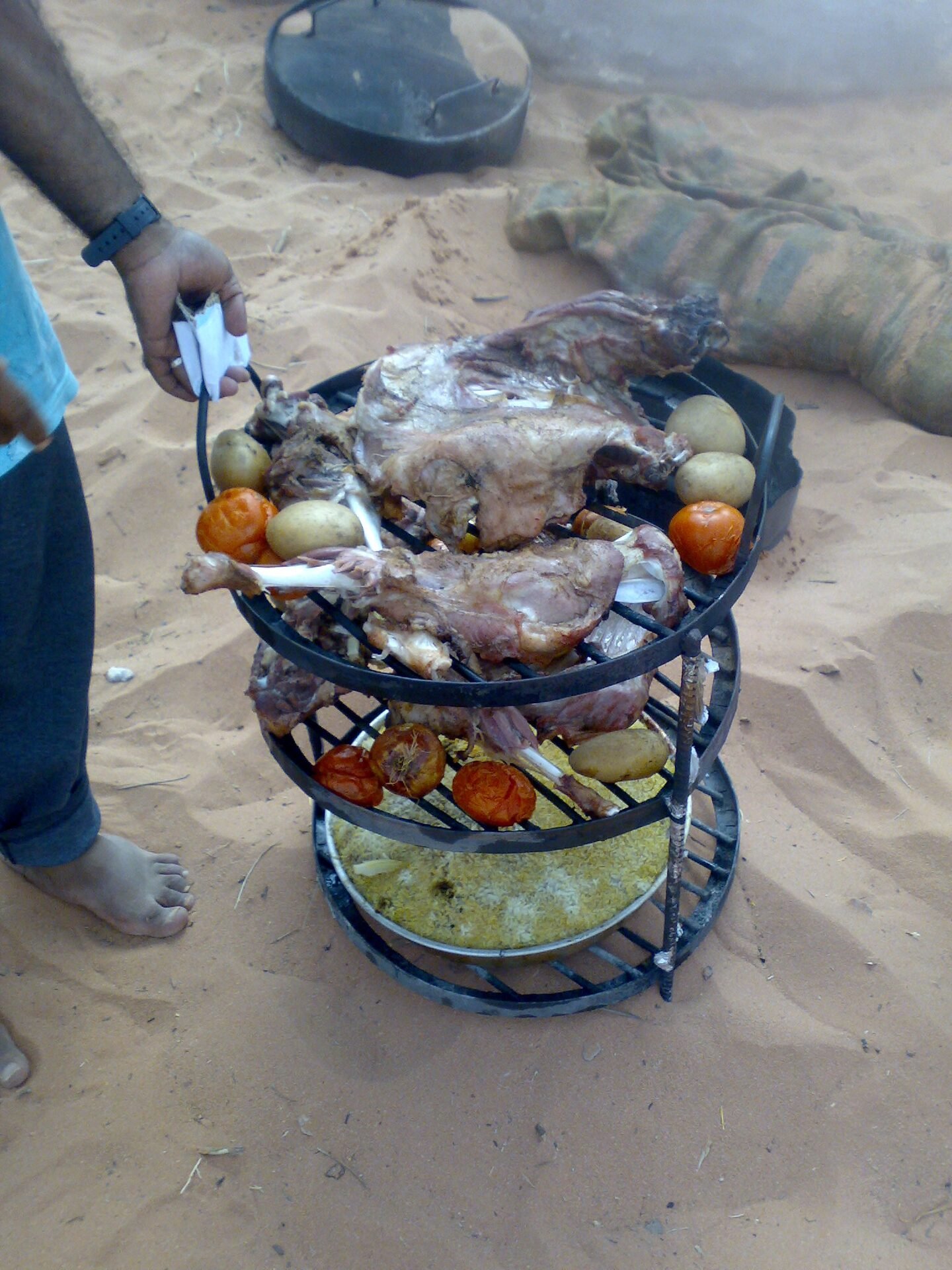 A tiered zarb rack lifted out of the sand oven, lamb on the top tier over tomatoes, potatoes and a tray of rice below.