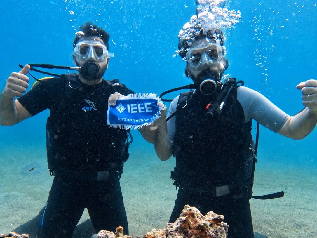 Two divers kneeling on the sand, one giving a thumbs-up, holding an IEEE Jordan Section pennant between them.