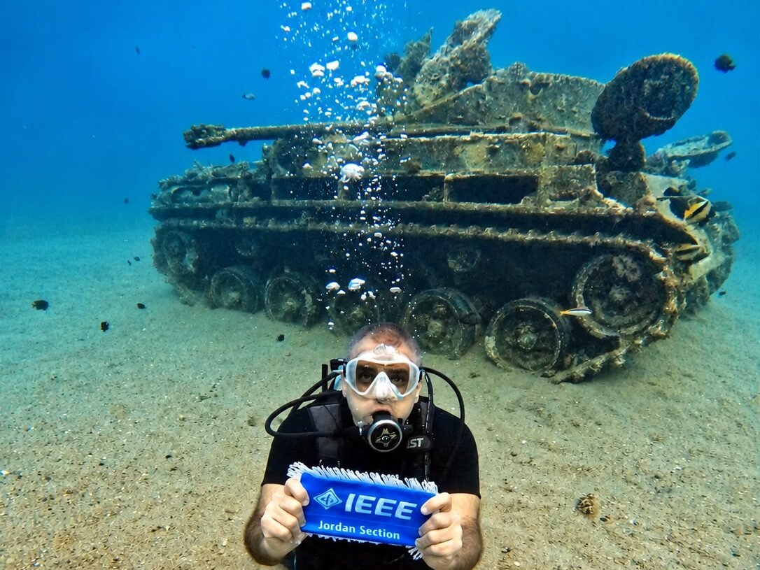 A diver kneeling on the sand in front of the sunken tank at Aqaba, holding an IEEE Jordan Section pennant up to the camera.