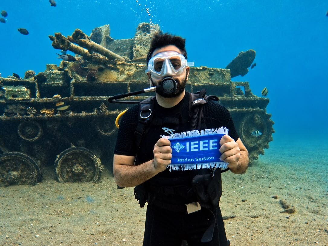 A diver kneeling on the sand beside the sunken tank, holding an IEEE Jordan Section pennant; the gun barrels and coral-covered tracks show behind.