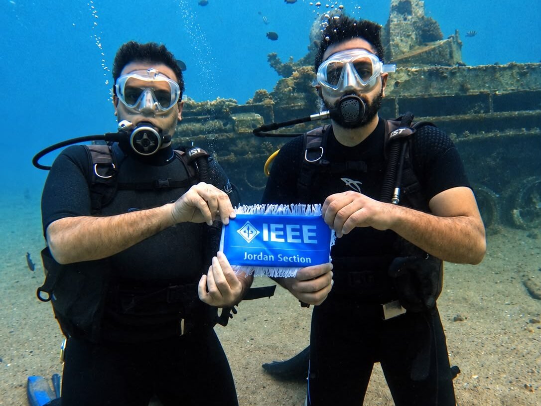 Two divers kneeling on the sand, holding an IEEE Jordan Section pennant between them, with the sunken tank behind.