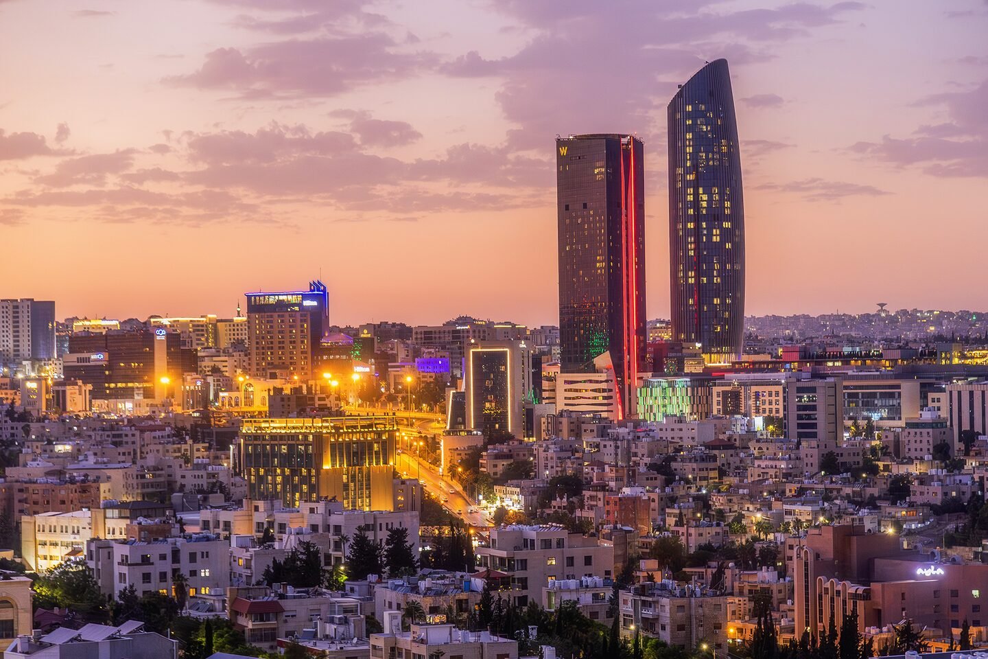 Amman skyline with lit high-rise towers at dusk under a violet and orange sky.